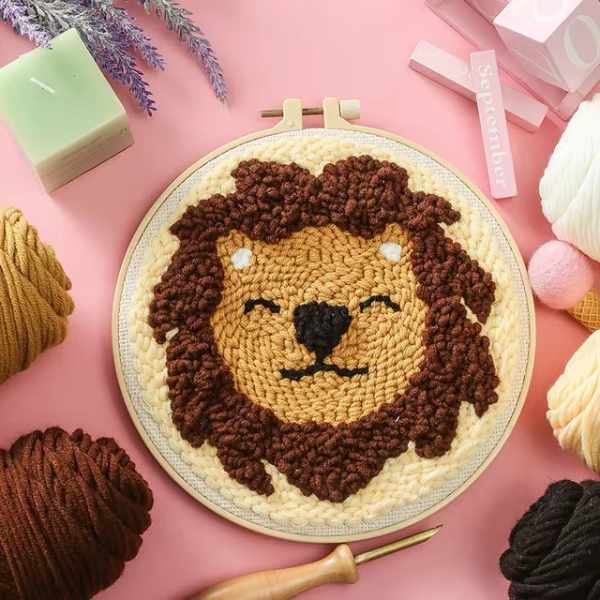 CraftHoop punch needle embroidery kit featuring a playful lion face design, displayed on a pink background with yarn and embroidery tools.