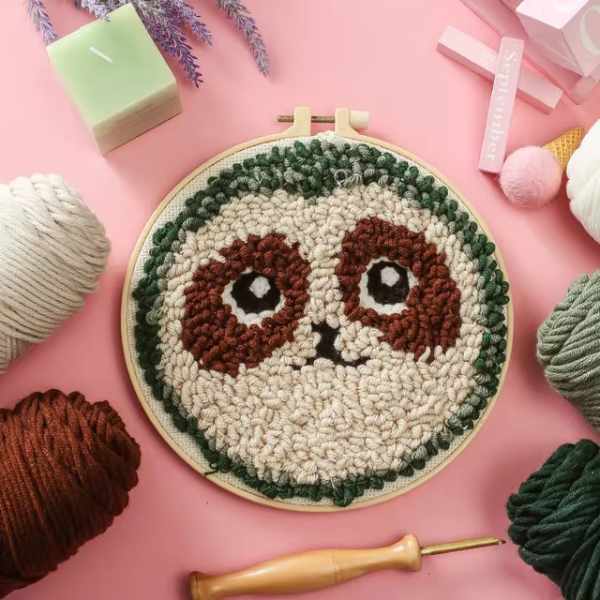 CraftHoop punch needle embroidery kit featuring an adorable sloth face design, displayed on a pink background with yarn and embroidery tools.