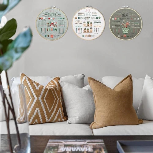 Three CraftHoop Beginner's 3-Piece Embroidery Kits with floral patterns hanging on a beige wall above a modern sofa with patterned pillows