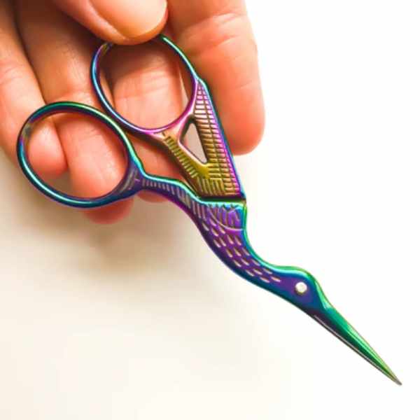 Close-up of rainbow-colored stork embroidery scissors, held between fingers, showcasing the elegant vintage design and detailed craftsmanship