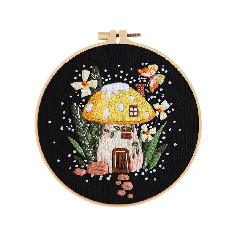 Close-up of CraftHoop mushroom embroidery kit with a yellow mushroom house design on black fabric, displayed on a wooden stand