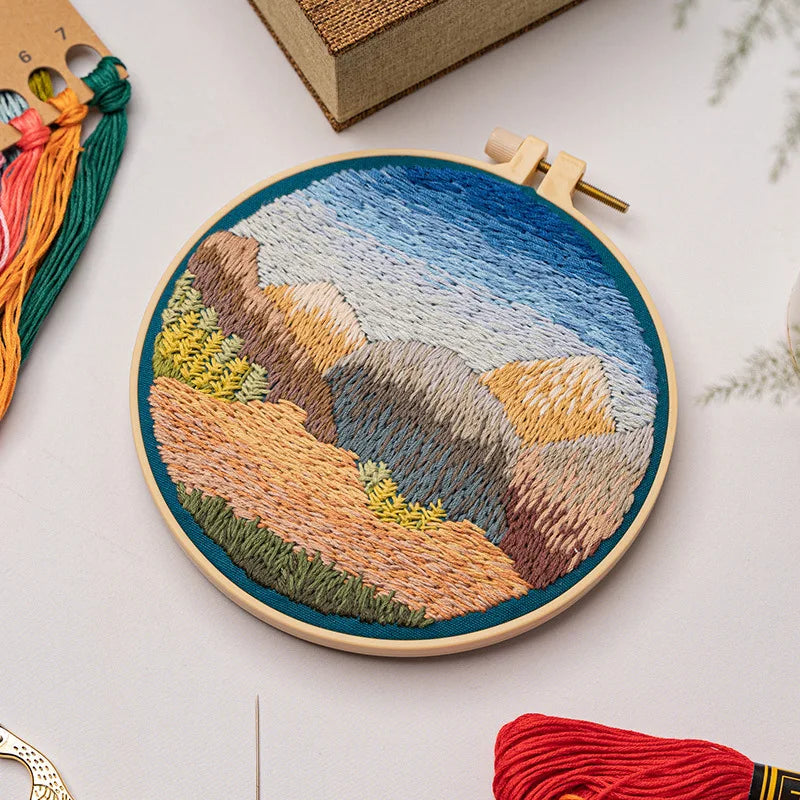 Close-up of CraftHoop landscape embroidery kit with a golden field and mountain design on fabric, displayed on a wooden surface with red and orange threads