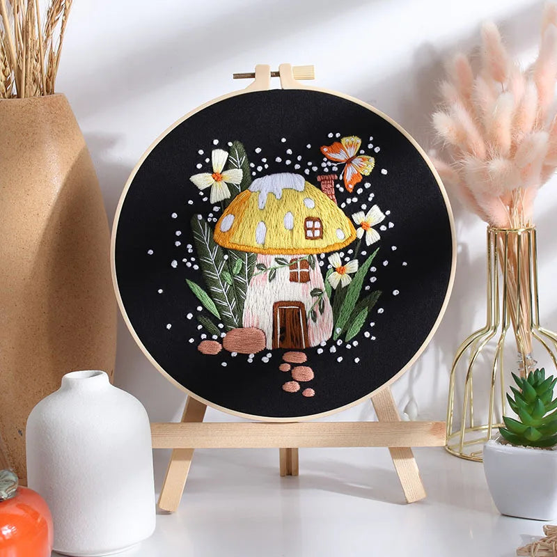 CraftHoop mushroom embroidery kit with red mushroom house design on black fabric, displayed on a wooden stand with decorative plants