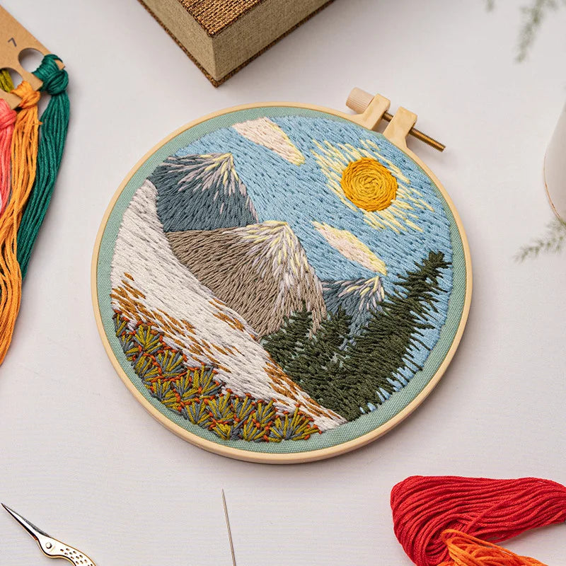 CraftHoop landscape embroidery kit showcasing a colorful sunset design with mountains and trees on fabric, displayed on a wooden surface with red and orange threads