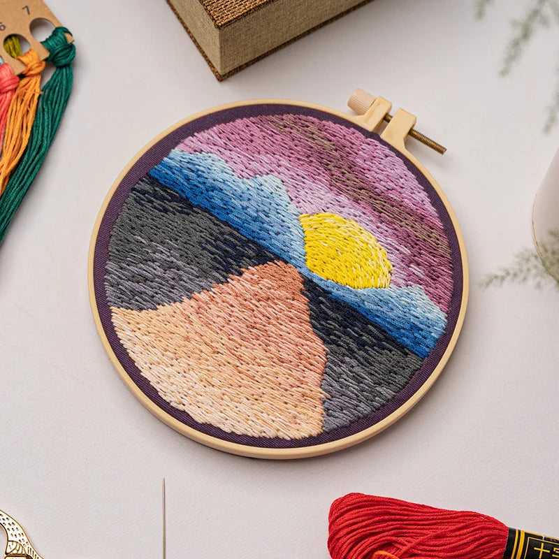 Close-up of CraftHoop landscape embroidery kit with a swirling river and mountain design on fabric, displayed on a wooden surface with embroidery tools