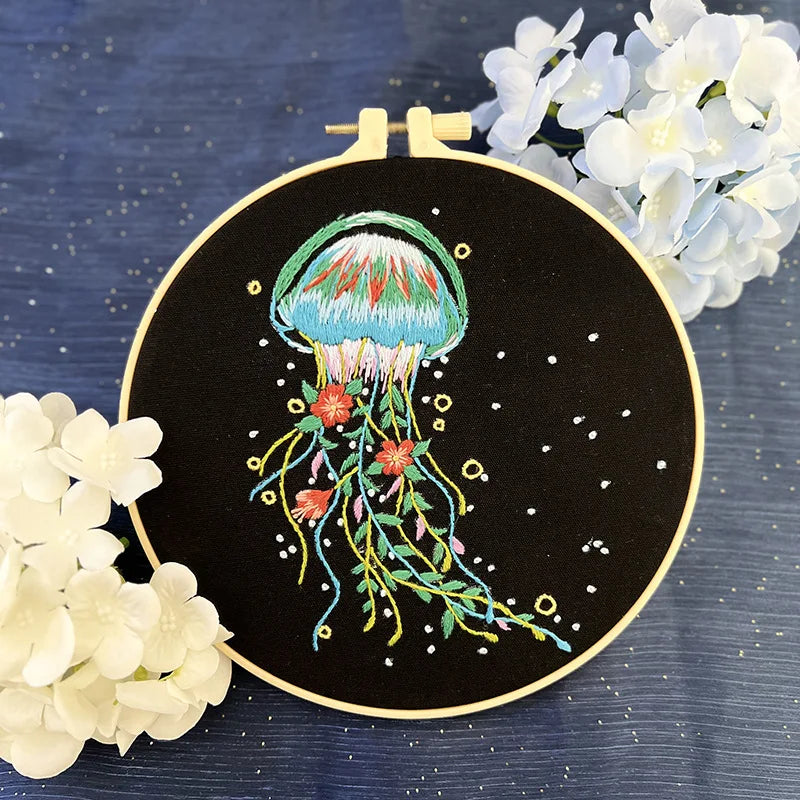 CraftHoop jellyfish embroidery kit with a pink and purple jellyfish design, featuring detailed tentacles on black fabric, displayed on a wooden stand with white flowers