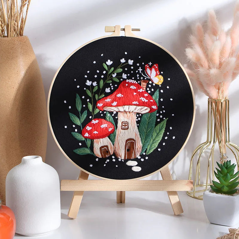 CraftHoop mushroom embroidery kit featuring a blue mushroom design on black fabric, displayed on a wooden stand with decorative elements