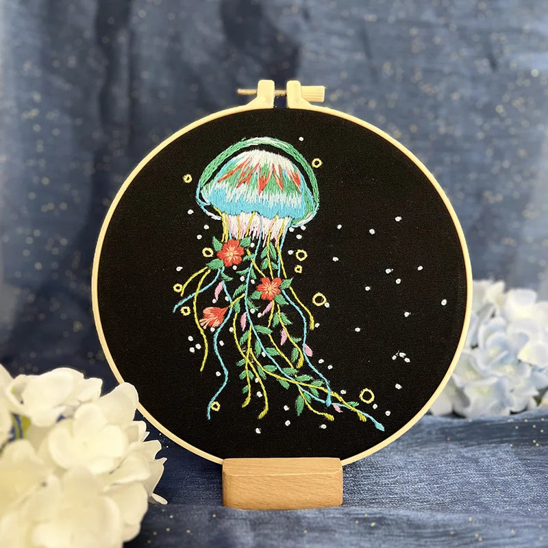 CraftHoop jellyfish embroidery kit with a green and yellow jellyfish design on black fabric, displayed on a wooden stand with white flowers