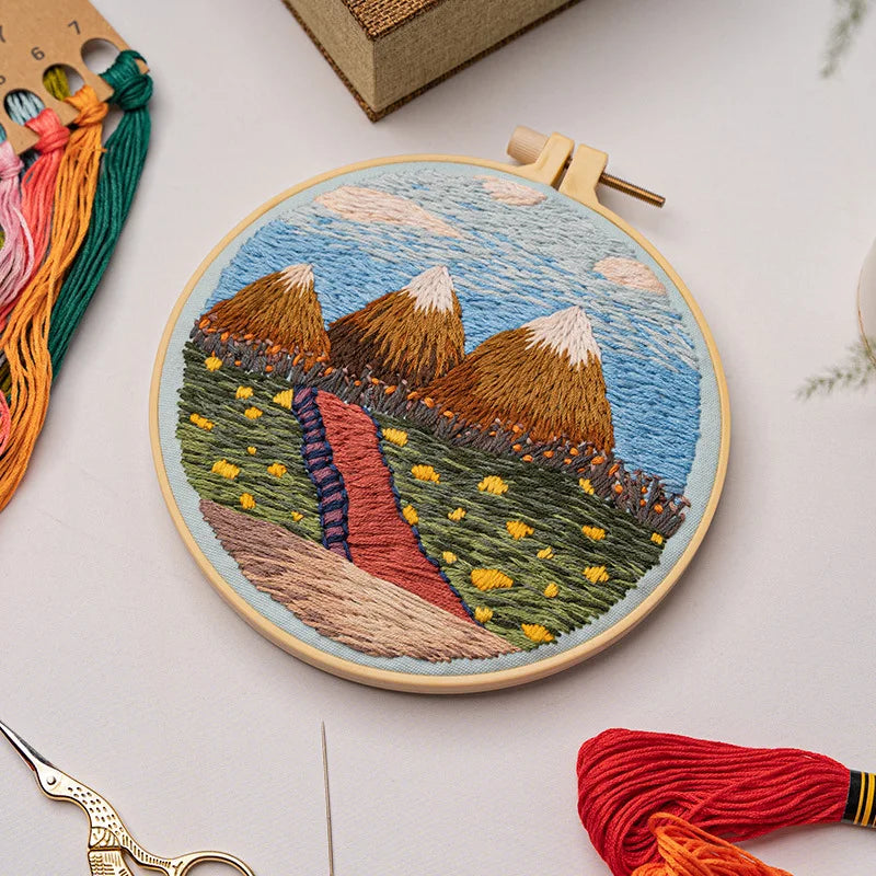 CraftHoop landscape embroidery kit featuring a colorful mountain scene with a sun design on fabric, displayed on a wooden surface with embroidery tools