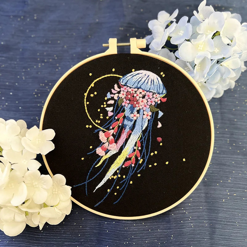 CraftHoop jellyfish embroidery kit featuring a multicolored jellyfish design with flowing tentacles on black fabric, displayed on a wooden stand with decorative sea elements