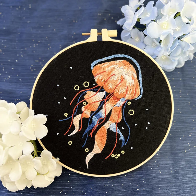 CraftHoop jellyfish embroidery kit with a pink and blue jellyfish design, showcasing fine details on black fabric, displayed on a wooden stand with white flowers