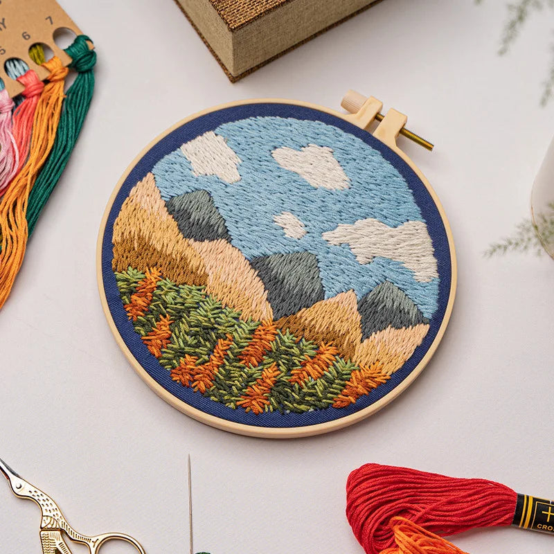 CraftHoop landscape embroidery kit with a farm and mountain scene, showcasing detailed stitching on fabric, displayed on a wooden surface with embroidery tools