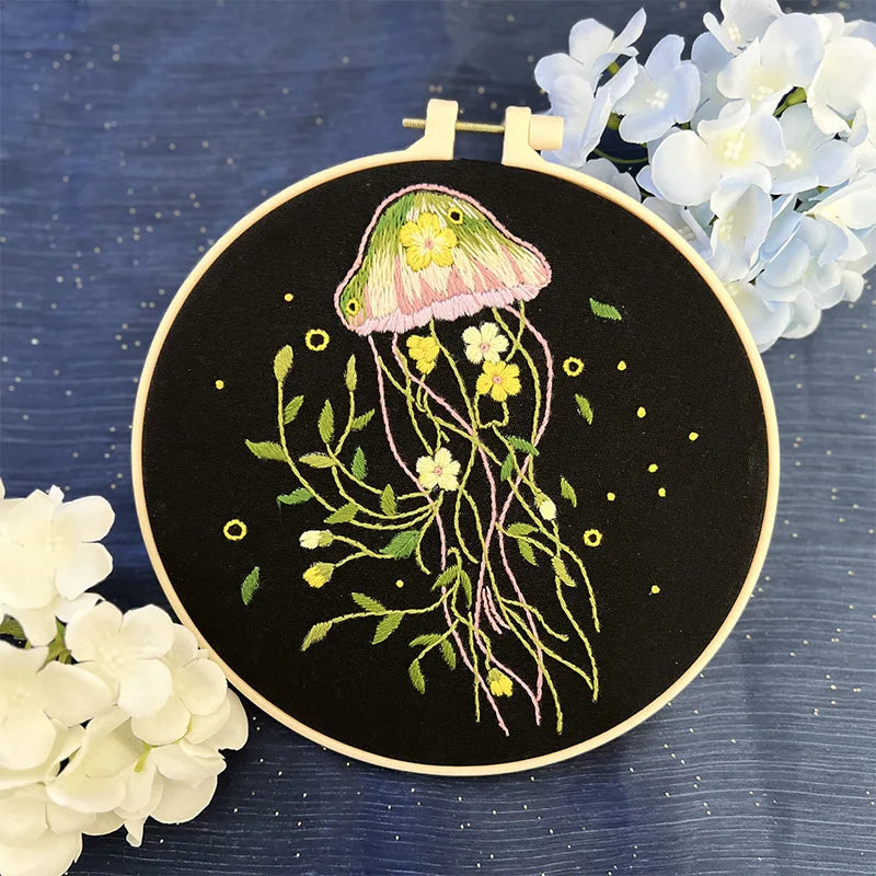 CraftHoop jellyfish embroidery kit featuring a green and yellow jellyfish design with intricate detailing on black fabric, displayed on a wooden stand with white flowers