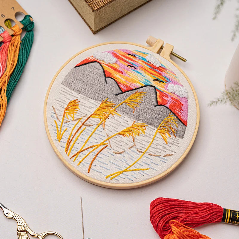 CraftHoop landscape embroidery kit showcasing a sunset over the mountains, with detailed stitching on fabric, displayed on a wooden surface