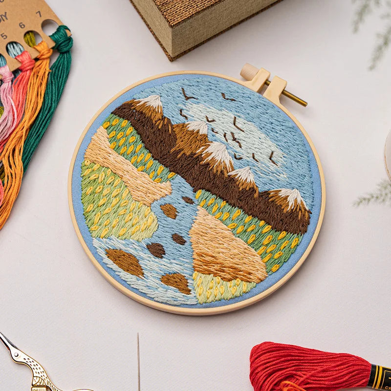 CraftHoop landscape embroidery kit featuring a peaceful river and mountain design on fabric, displayed on a wooden surface with red and orange threads