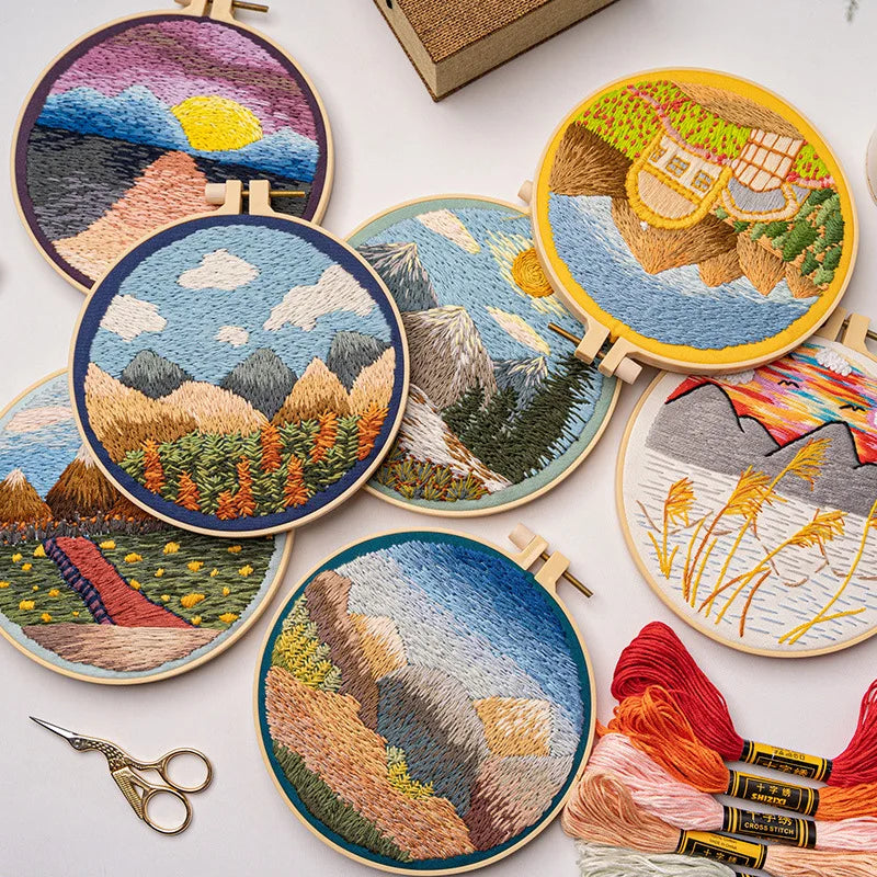 CraftHoop embroidery kit featuring a scenic mountain landscape with trees and sun design on blue fabric, displayed on a wooden surface with red and orange threads