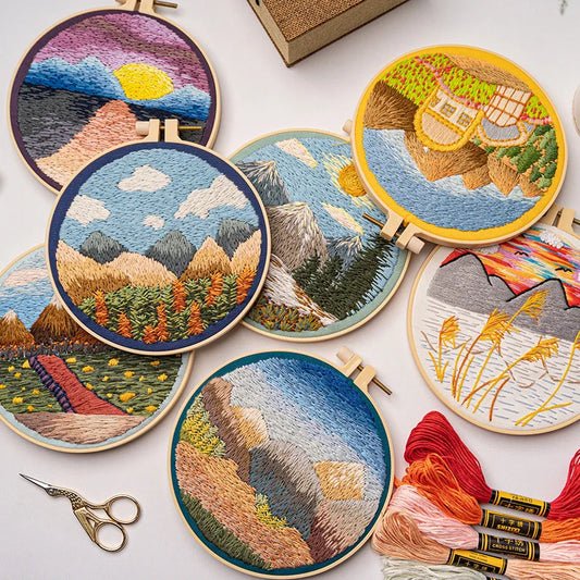 CraftHoop embroidery kit featuring a scenic mountain landscape with trees and sun design on blue fabric, displayed on a wooden surface with red and orange threads